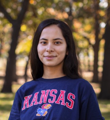 Aylar, wearing a Kansas sweatshirt and standing in front of several trees
