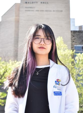 A woman in a white lab coat in front of a building and trees